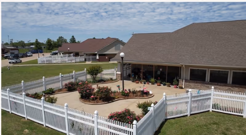 Exterior view of a white-picket-fenced courtyard with flower beds and potted plants beside a single-story facility building.