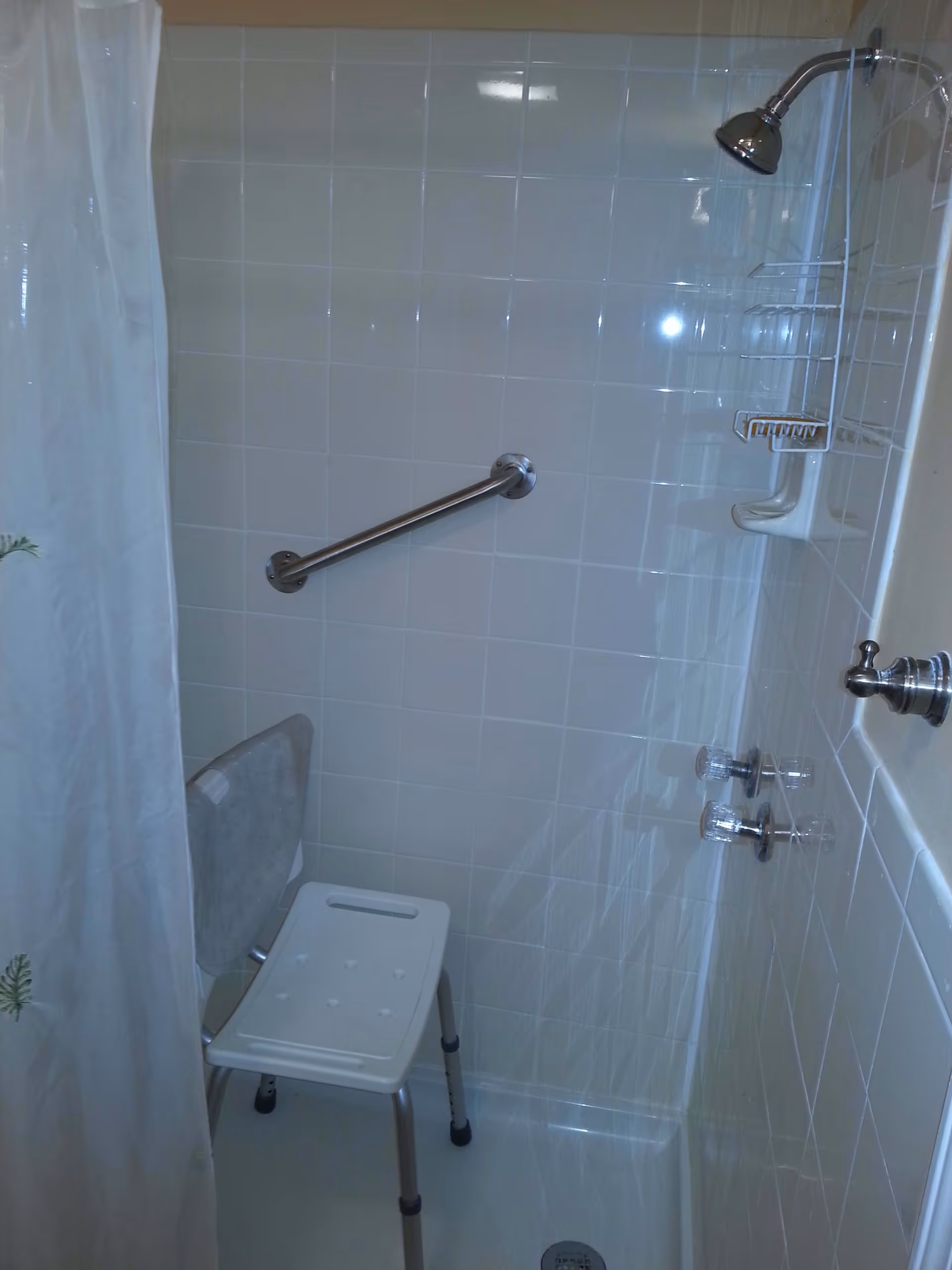 A tiled shower area with a white shower chair, a metal grab bar on the wall, a showerhead, and a shower curtain partially visible on the left side.