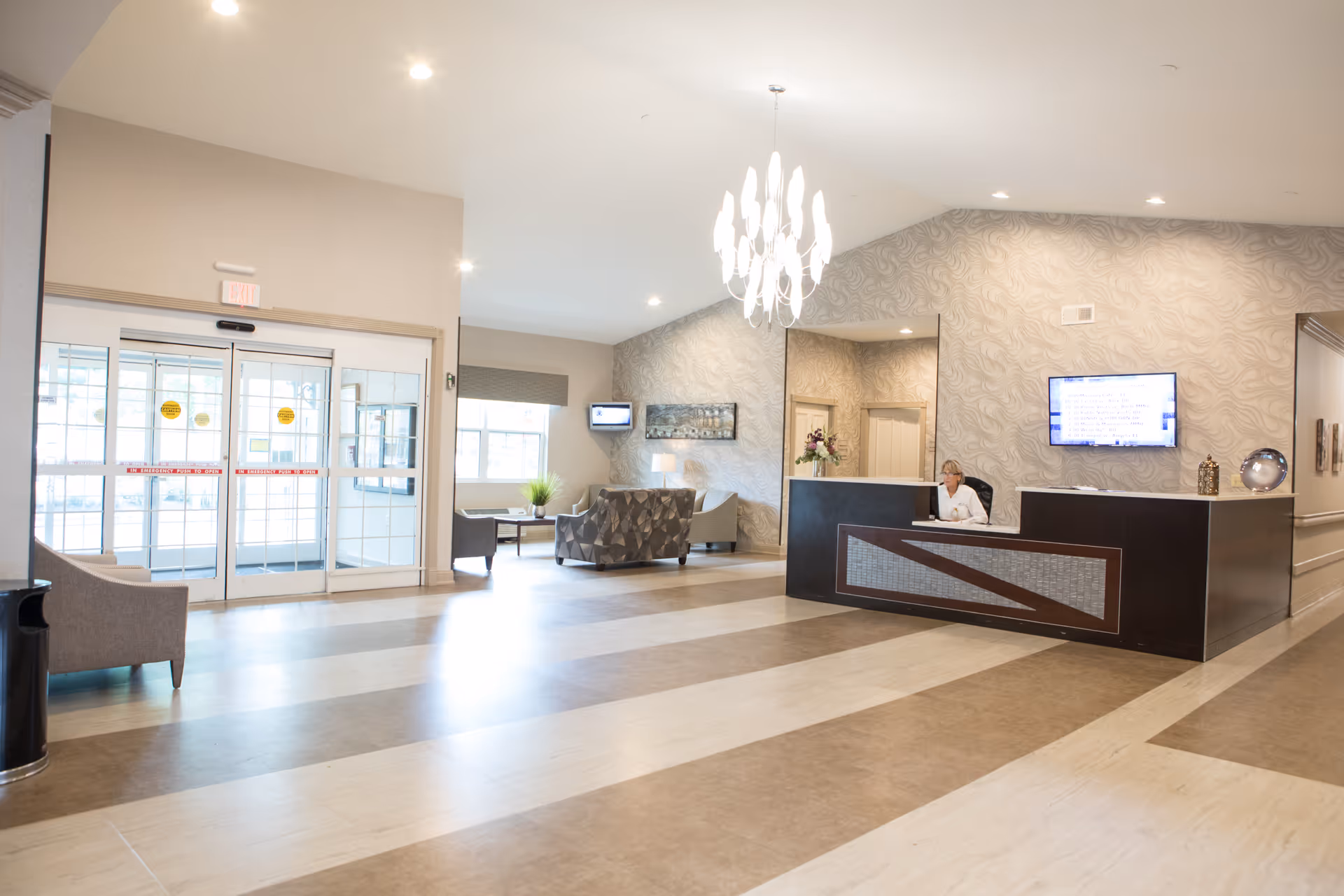 A spacious and well-lit assisted living facility lobby with a reception desk where a staff member is seated. The lobby features modern decor with patterned wallpaper, a chandelier, seating area with chairs and a sofa, large windows, and automatic glass entrance doors.