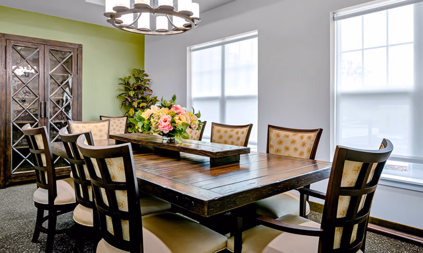 A dining room with a large wooden table surrounded by eight cushioned chairs. The table has a decorative centerpiece with a bouquet of flowers. There is a green accent wall with a tall cabinet featuring glass doors and a leafy plant next to it. Two large windows with white blinds allow natural light into the room. A modern chandelier hangs above the table.
