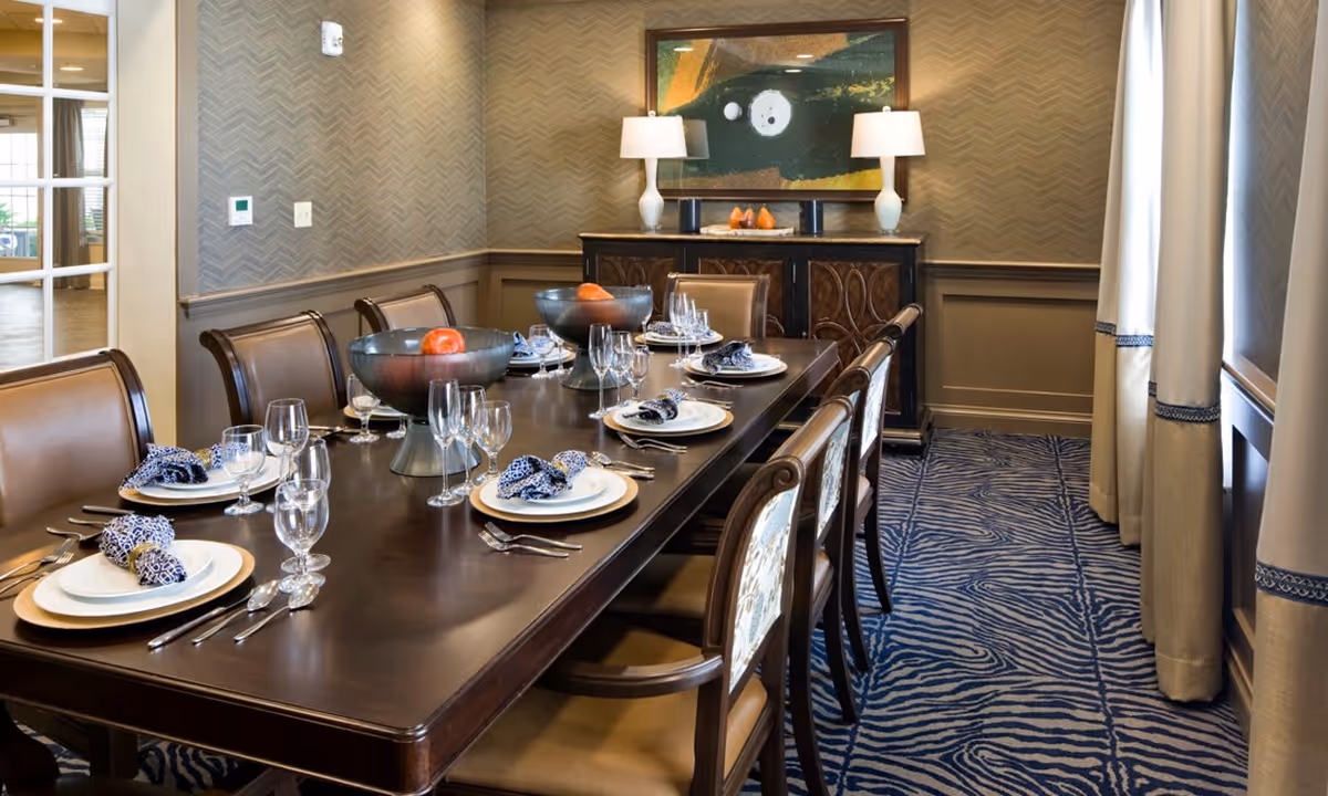 A formal dining room with a long dark wooden table set for eight people. Each place setting includes a white plate, silverware, a water glass, a wine glass, and a blue patterned cloth napkin. Two large decorative bowls with fruit are placed on the table. The room features patterned wallpaper, a blue and white zebra-striped carpet, beige curtains with blue trim, and a sideboard with two lamps and a framed abstract painting above it.