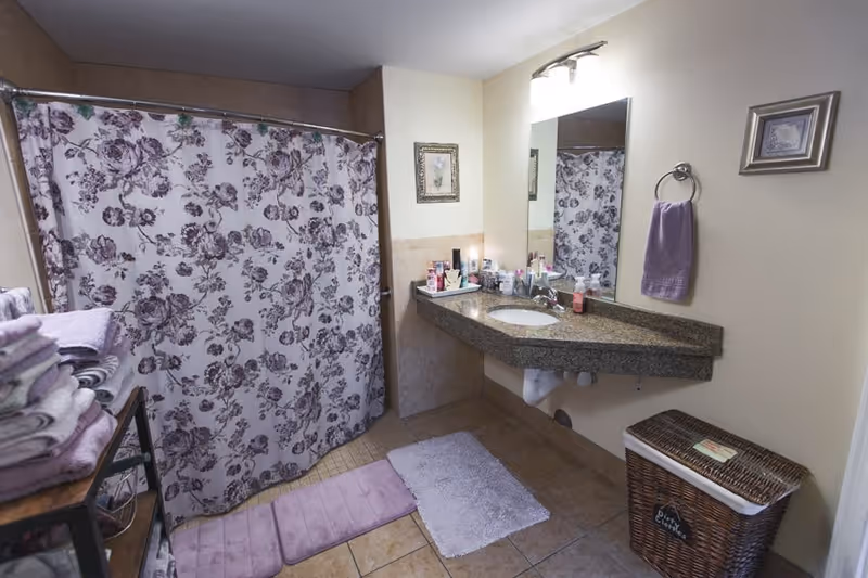 Bathroom with a floral shower curtain, granite countertop sink, folded towels, bath mats, and a wicker laundry basket.