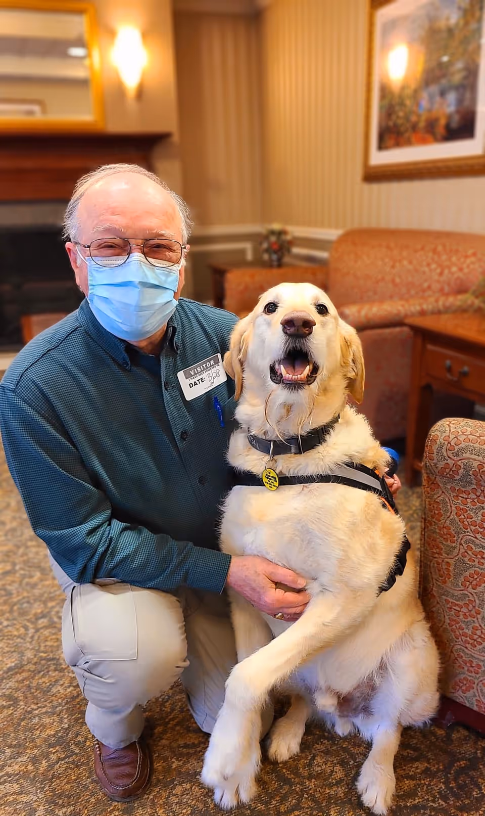 An elderly man wearing glasses and a face mask kneels on the carpeted floor of a cozy living room area, smiling and holding a large golden retriever dog that is sitting beside him. The room has warm lighting, patterned furniture, a framed picture on the wall, and a fireplace in the background.