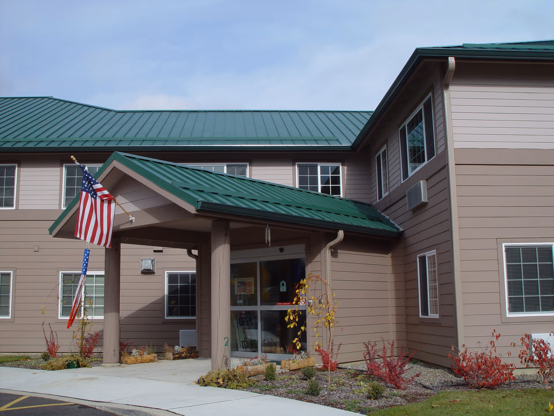 Front entrance of a two-story building with a green metal roof, American flag, and glass double doors.