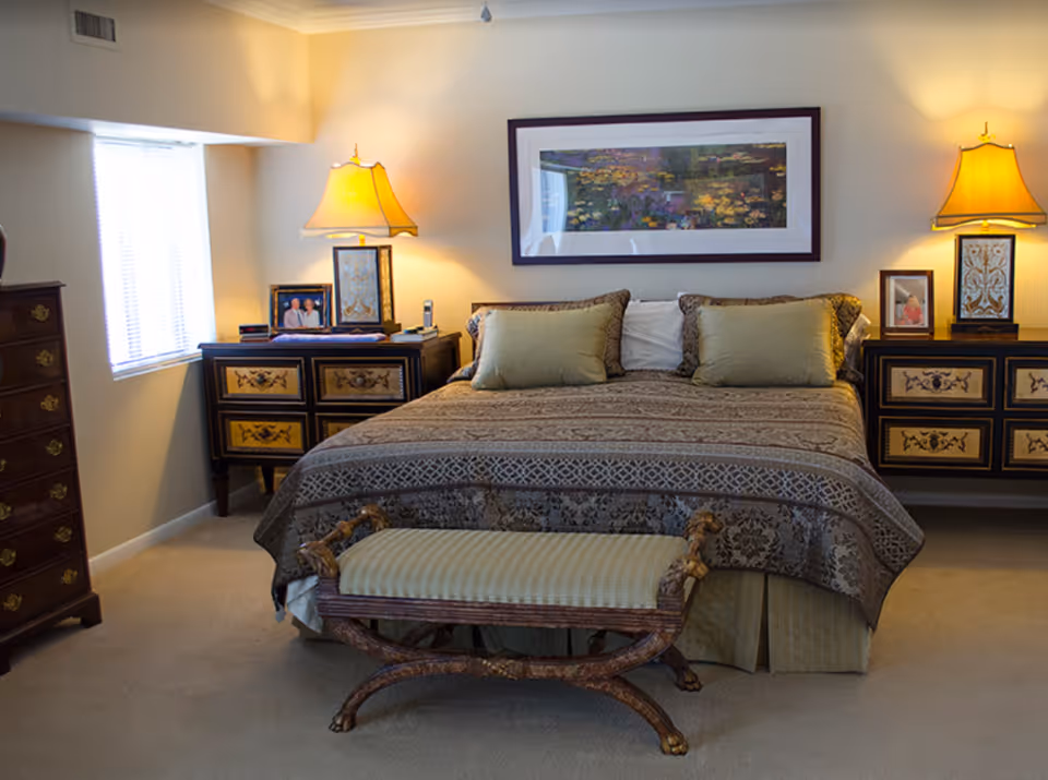 A well-decorated bedroom featuring a large bed with patterned bedding and two green pillows. On either side of the bed are matching nightstands with ornate designs, each holding a lamp and framed photographs. A cushioned bench with wooden legs is placed at the foot of the bed. A large framed artwork hangs on the wall above the bed, and a window with blinds is visible on the left side of the room.