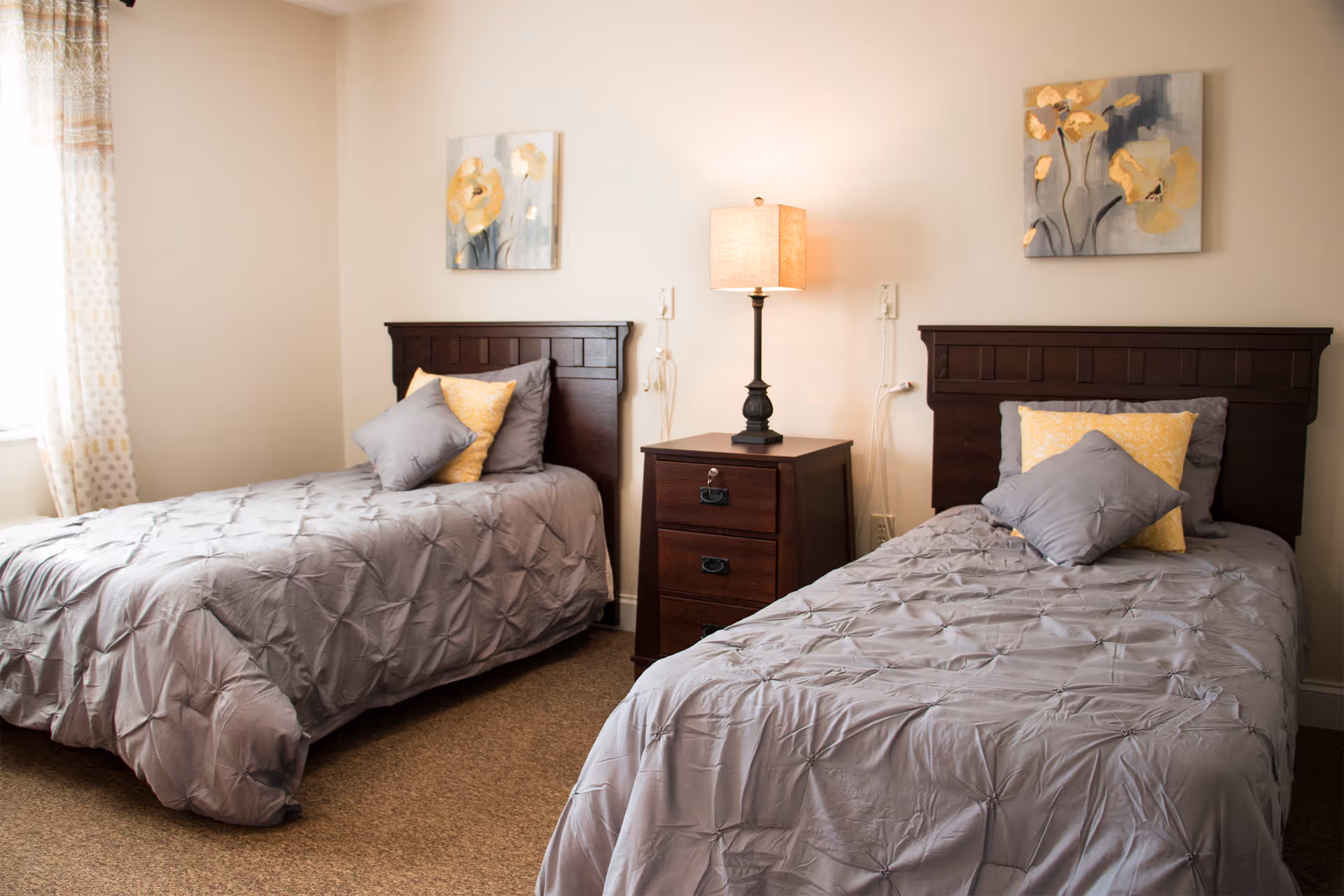 A tidy bedroom with two twin beds dressed in gray bedding with yellow accent pillows, a central nightstand and lamp, and floral wall art.