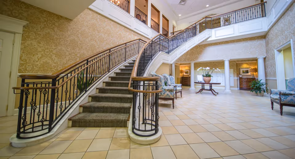 Spacious and elegant interior of a senior living facility featuring a wide staircase with ornate black railings and wooden handrails. The floor is tiled with light-colored tiles, and the walls are decorated with patterned wallpaper. There are several upholstered armchairs with floral patterns arranged around the room, a round wooden table with a large potted plant in the center, and two white columns supporting the upper floor. The area is well-lit with natural light coming from windows above.