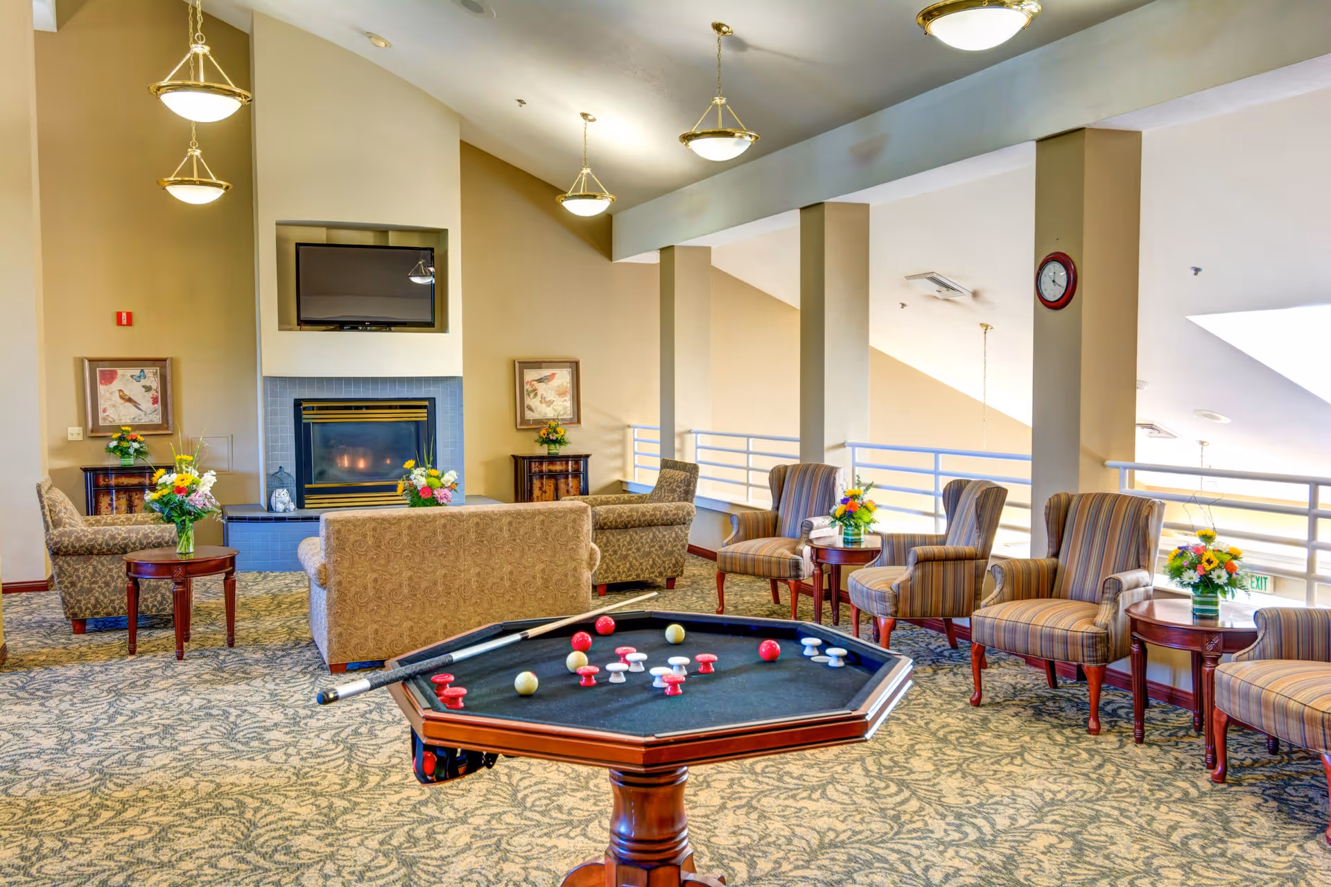 A spacious and well-lit living room area in an assisted living facility featuring comfortable upholstered chairs and sofas arranged around a fireplace with a mounted TV above it. There are side tables with flower arrangements, hanging ceiling lights, and a game table with colorful game pieces in the foreground. The room has a patterned carpet and large windows allowing natural light.