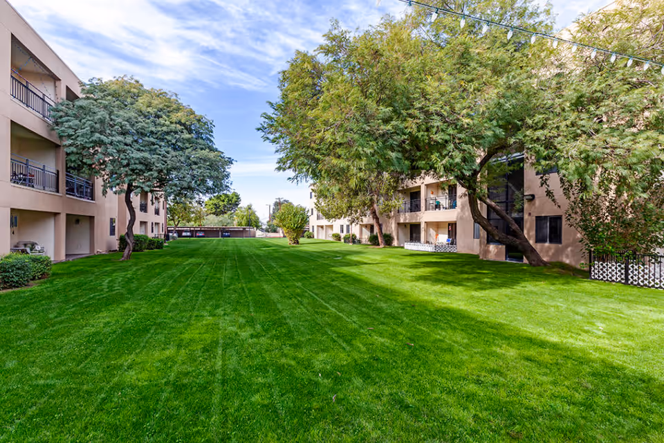 A well-maintained green lawn area between two beige multi-story residential buildings with balconies. Several mature trees provide shade, and the sky is partly cloudy with blue patches.
