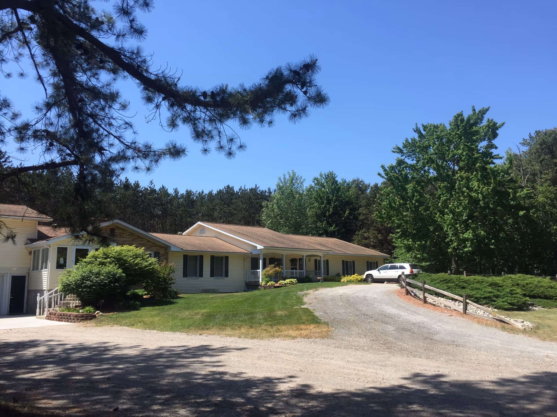 A single-story residential building with a light yellow exterior and a brown roof, surrounded by green trees and bushes under a clear blue sky. A white car is parked on a gravel driveway leading to the building, which has a small porch with white railings.