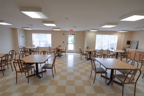 A bright dining room with several square tables and wooden chairs arranged neatly. The room has large windows letting in natural light, beige and white checkered flooring, and a door at the far end leading outside. The walls are light-colored, and there is a cabinet with a framed sign on top along the right side.