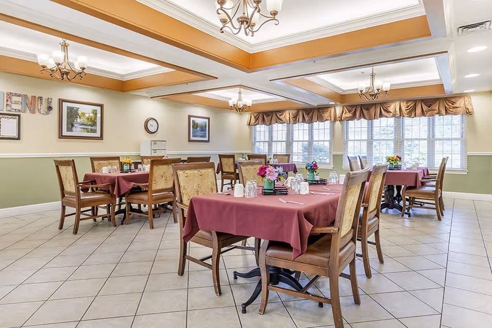 A bright dining room with several wooden tables covered with burgundy tablecloths, each set with glasses, cups, and small flower arrangements. The room has large windows with brown valances, tiled floors, and chandeliers hanging from a coffered ceiling. Framed pictures and a clock are on the walls.