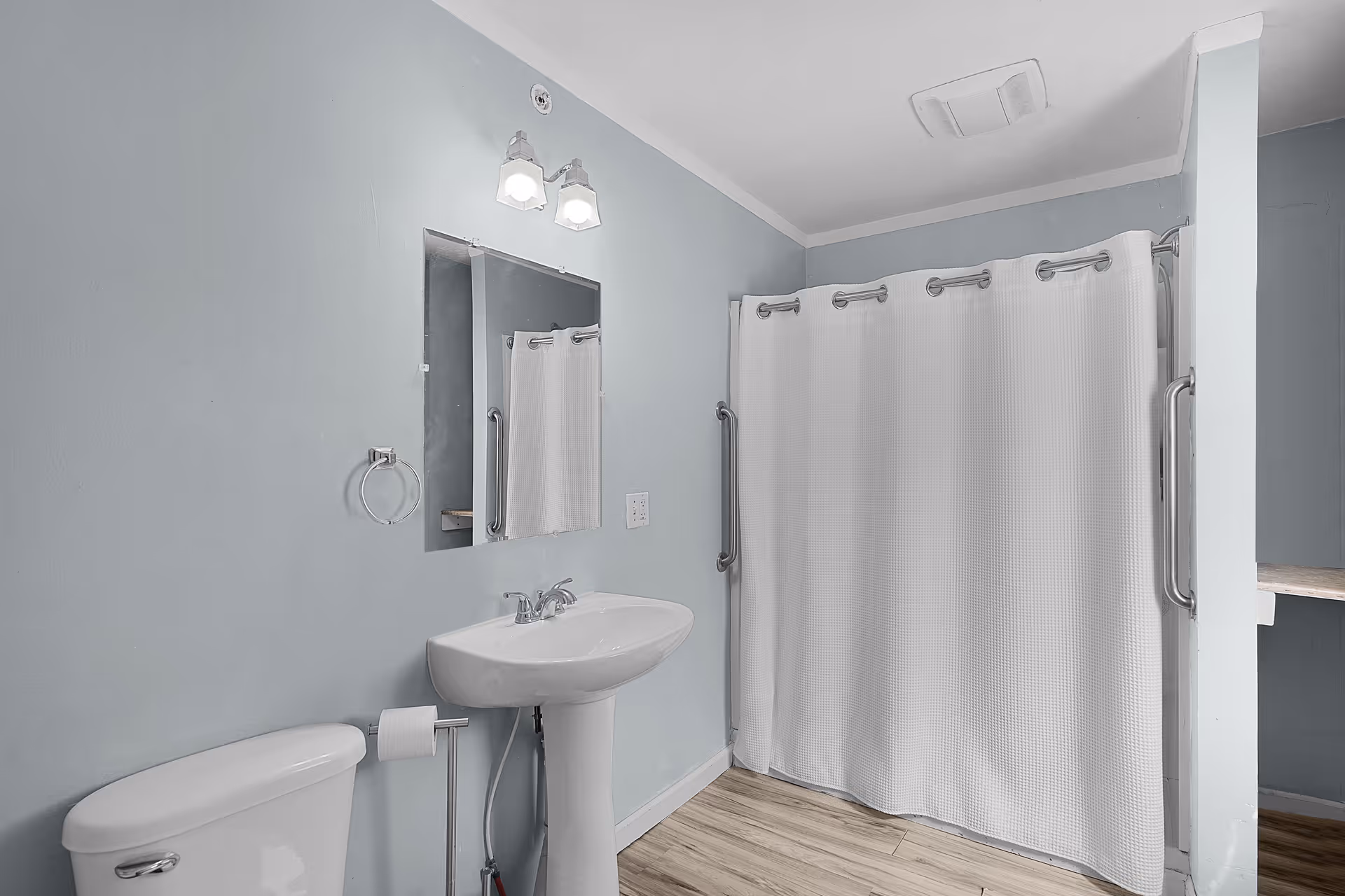 A clean bathroom with light blue walls featuring a white toilet, a pedestal sink with a mirror above it, a towel ring, and a shower area with a white curtain and grab bars on both sides. The floor is wood-patterned.
