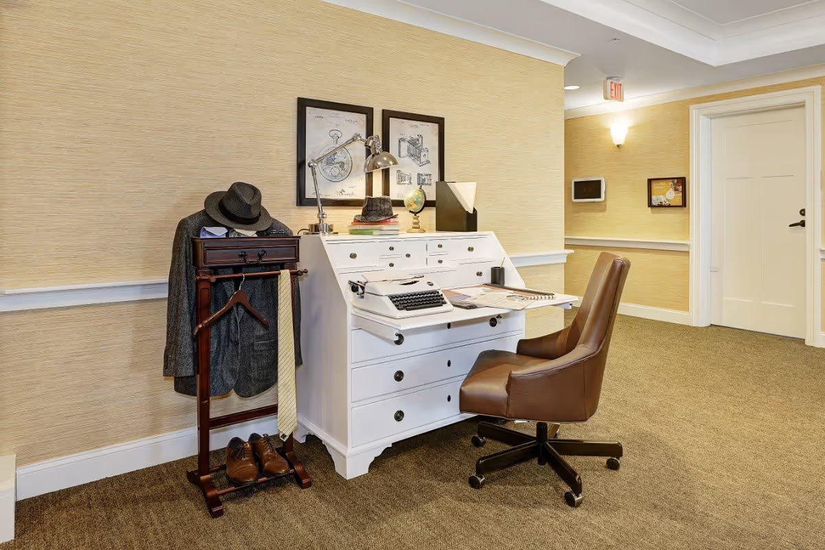 A cozy office corner with a white vintage-style desk featuring a typewriter, books, a globe, and a desk lamp. Next to the desk is a wooden valet stand holding a gray jacket, a hat, a tie, and a pair of brown shoes. A brown leather office chair is positioned in front of the desk. The walls are covered with light beige wallpaper, and two framed technical drawings hang above the desk. The carpet is brown, and a white door and hallway are visible in the background.