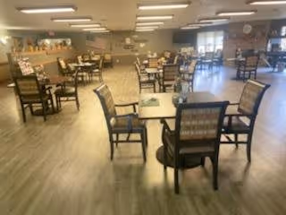 A spacious dining room with multiple wooden tables and chairs arranged neatly on a light wood floor. The room is well-lit with ceiling lights and has large windows allowing natural light to enter. The walls are decorated with bulletin boards and some artwork.