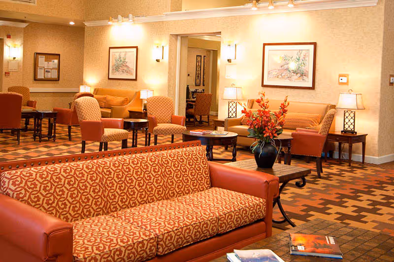A warm and inviting senior living facility common area with patterned orange and beige sofas and armchairs, small tables with lamps, framed artwork on the walls, and a vase with orange flowers on a coffee table. The room has a patterned carpet and soft lighting.