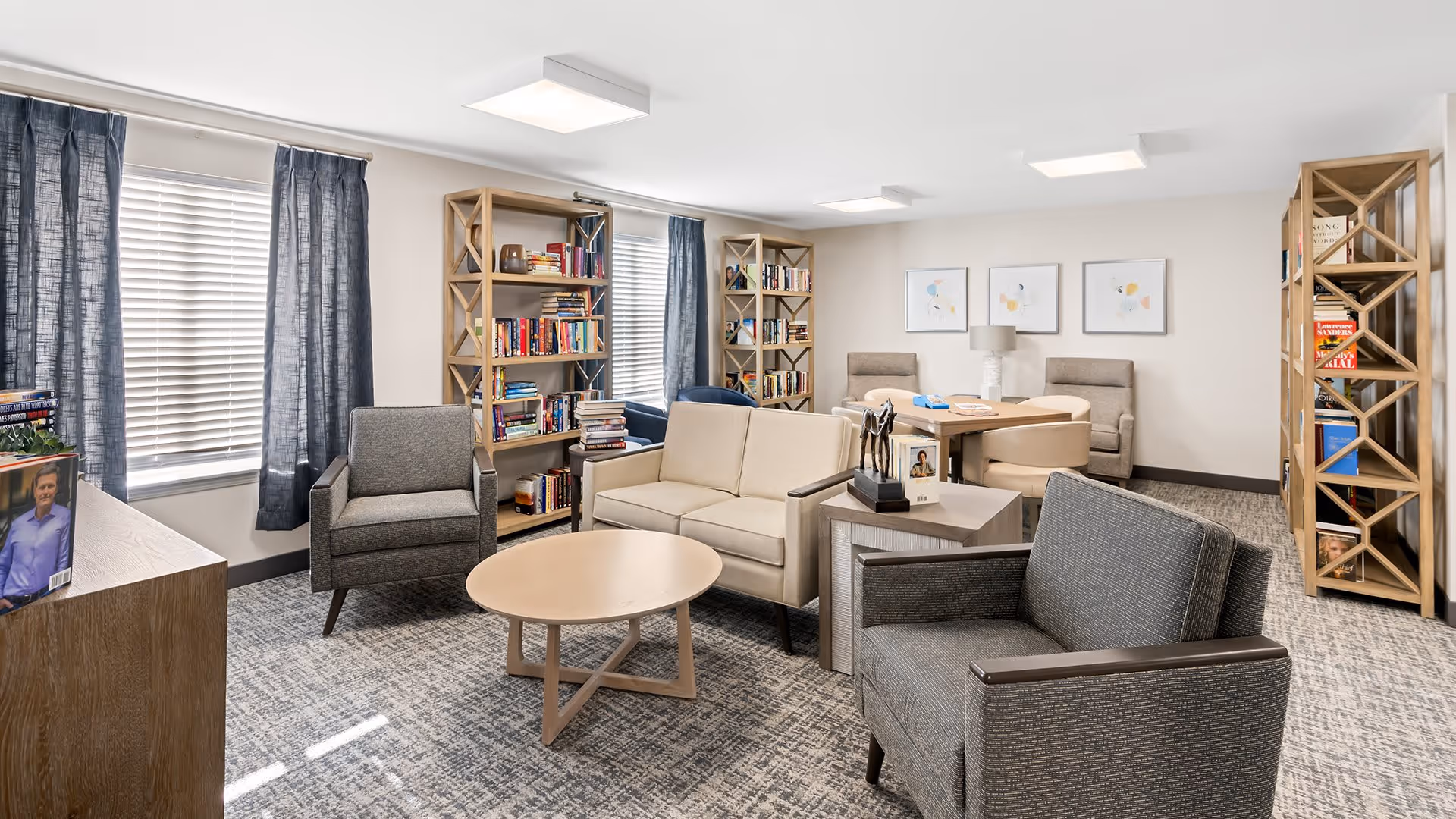 A bright and cozy living room area in a senior living facility with beige and gray armchairs, a beige loveseat, a round wooden coffee table, and multiple wooden bookshelves filled with books. The room has large windows with blue curtains and white blinds, light-colored walls, and framed abstract artwork on the far wall. There is also a small table with chairs and a lamp in the background.