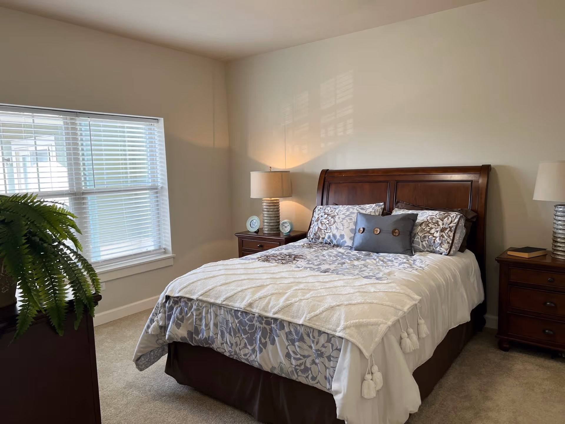 A neatly made bed with floral patterned bedding and decorative pillows in a bedroom with beige walls. There are two wooden nightstands on either side of the bed, each with a lamp. A window with white blinds is on the left side, and a green potted plant is partially visible in the foreground.