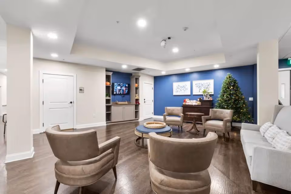 A modern living room with a blue accent wall, decorated with two framed artworks and a Christmas tree. The room features a light gray sofa with pillows, four beige armchairs arranged around a round coffee table, and a wooden cabinet with decorative items. The floor is dark wood, and the ceiling has recessed lighting.