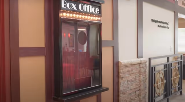 Interior view of a senior living facility hallway featuring a black box office ticket window with illuminated lights above it. The wall has a combination of beige and brown panels with a stone accent and decorative metal railing on the right side.