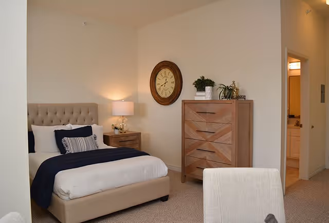 Well-lit bedroom with a tufted bed, nightstand and lamp, a wooden dresser with decor, and a doorway leading to a bathroom.