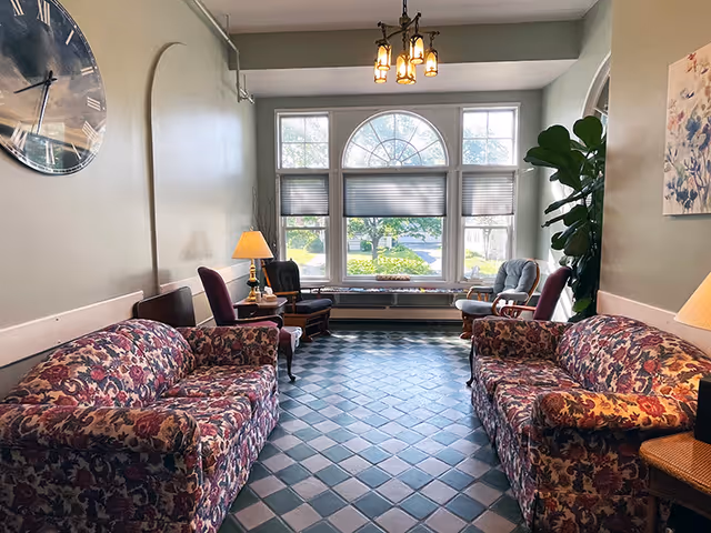 A cozy sitting area with two floral patterned sofas facing each other on a tiled floor. There are several chairs and a table with a lamp near a large window with an arched top, allowing natural light to brighten the room. A large wall clock and a potted plant are also visible.