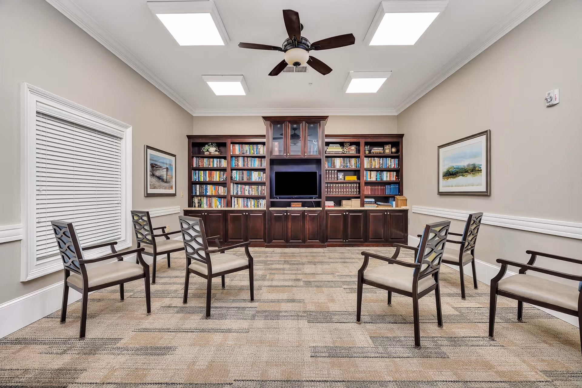 A quiet room with six chairs arranged in a circle on a patterned carpet. The back wall features a large dark wood bookshelf filled with books and a television in the center. The walls are painted light beige with white trim, and two framed landscape paintings hang on either side. The ceiling has recessed lighting and a ceiling fan with dark blades.