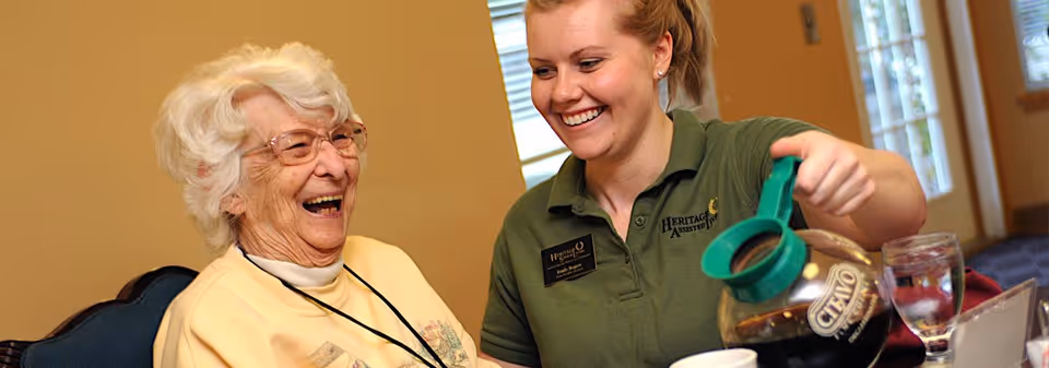 A smiling caregiver pours coffee for a laughing elderly woman seated at a dining table inside a senior living facility.