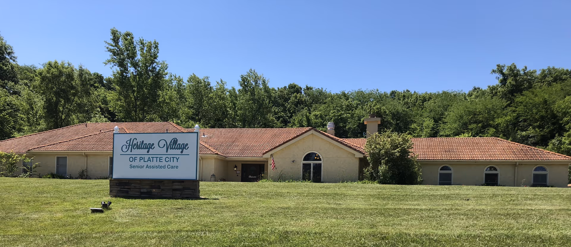 Single-story senior assisted living building with a tiled roof, front lawn, and a sign reading 'Heritage Village of Platte City Senior Assisted Care'.
