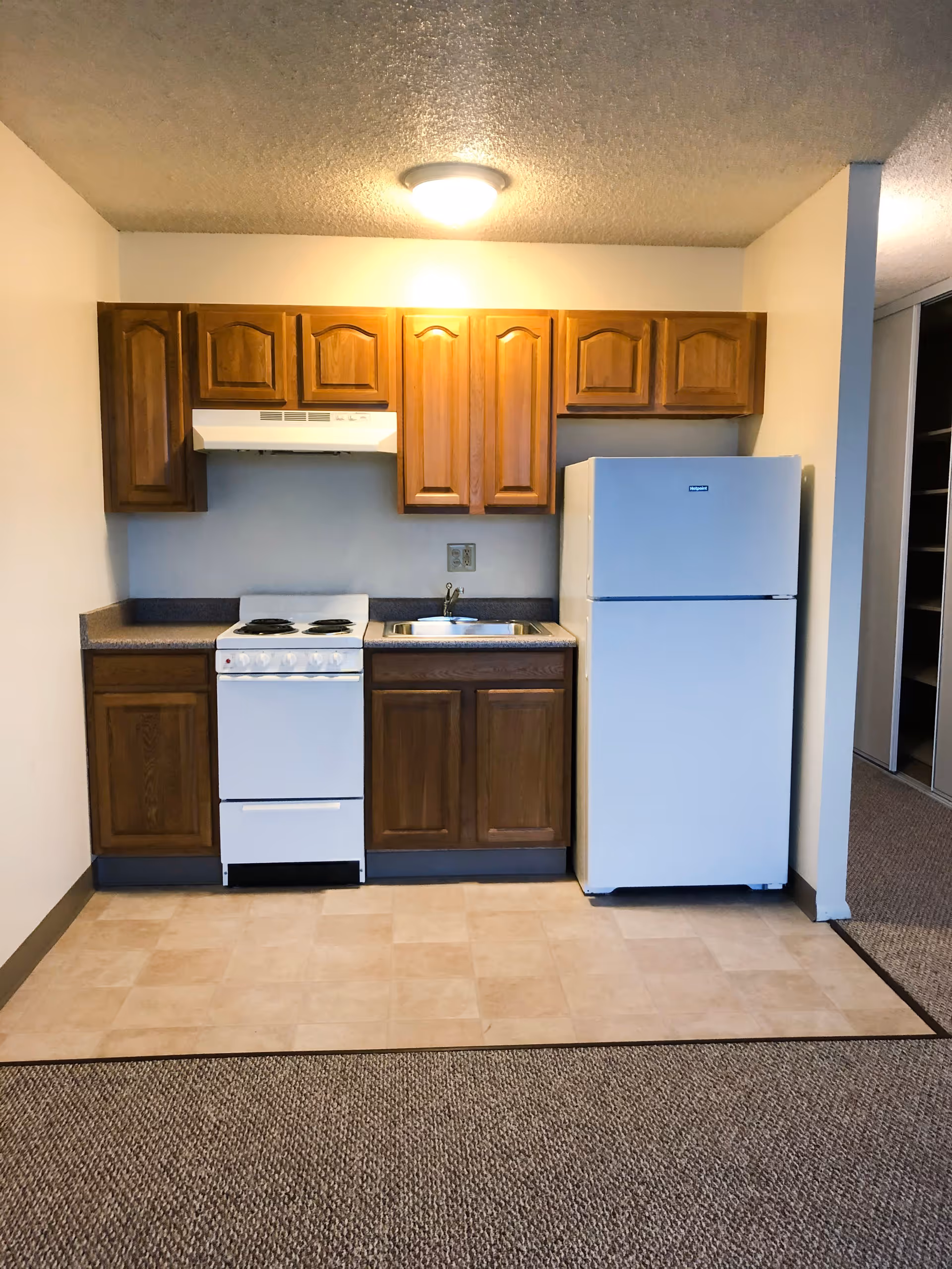 Small kitchen area with wooden cabinets, a white stove with four burners, a stainless steel sink, and a white refrigerator. The floor in front of the kitchen is tiled, transitioning to carpet in the adjacent area.