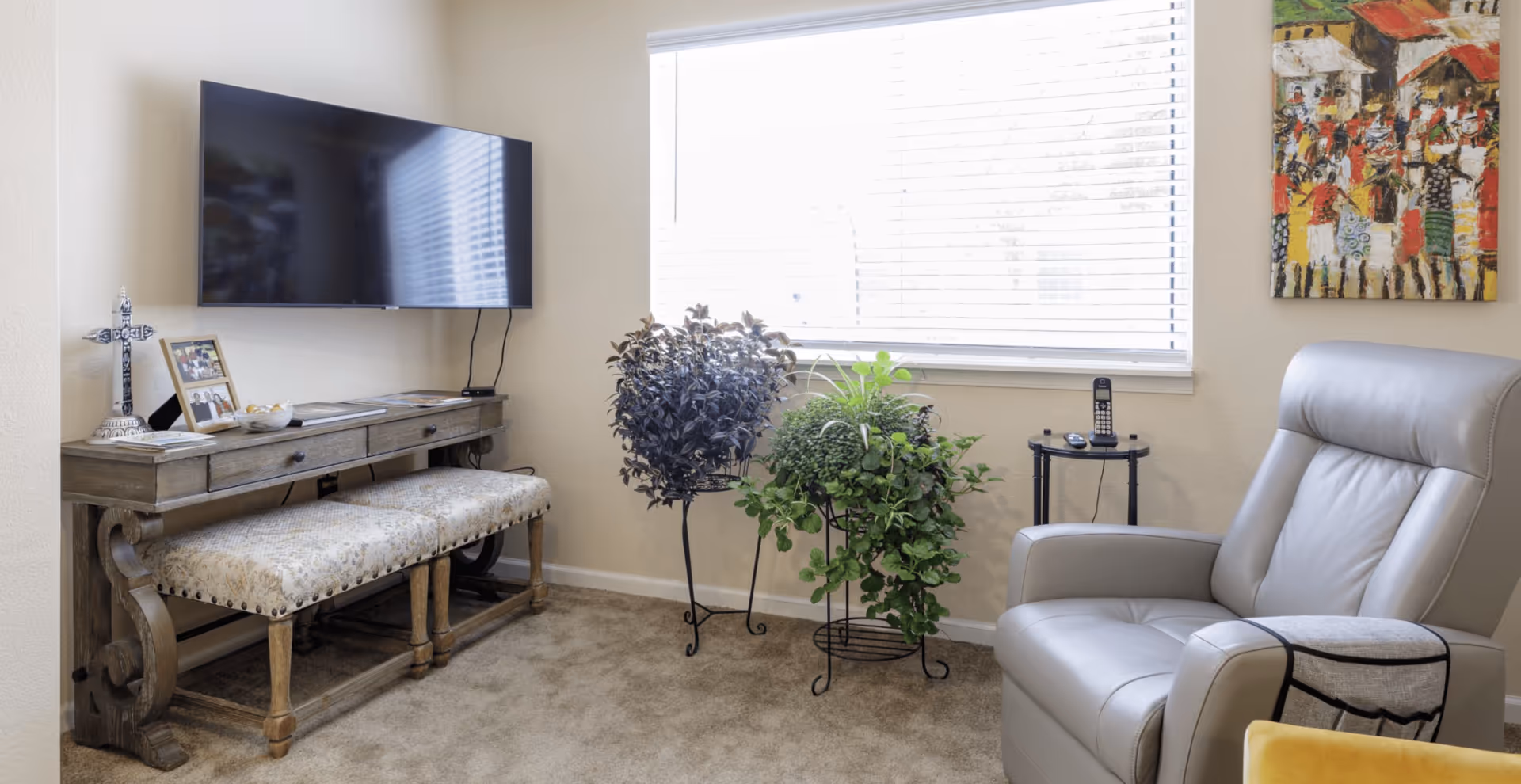 A cozy living room area with a mounted flat-screen TV above a wooden console table. The table has decorative items including a cross, framed photos, and a bowl. Below the table is a cushioned bench with a patterned fabric. To the right, there are two potted plants on black metal stands in front of a window with white blinds. A comfortable gray leather recliner chair is positioned near a small round side table holding a cordless phone and remote controls. A colorful painting hangs on the wall above the chair.