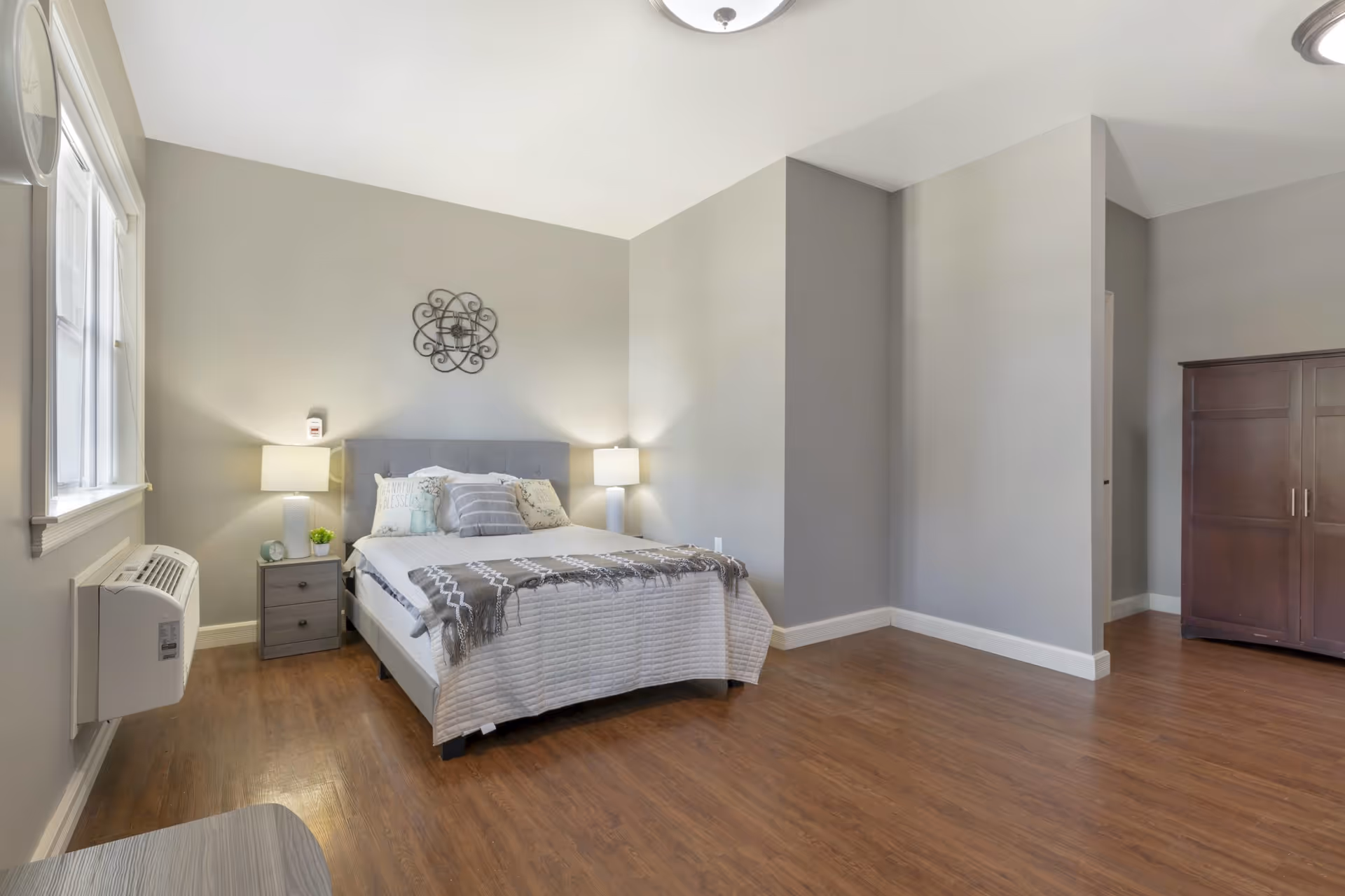 A clean and spacious bedroom with a bed centered against a light gray wall. The bed has a gray upholstered headboard, white bedding, and decorative pillows. There are two bedside tables with lamps on each side of the bed. A decorative wall piece hangs above the bed. The room has wood flooring, a large window on the left, and a dark wooden wardrobe on the right side.