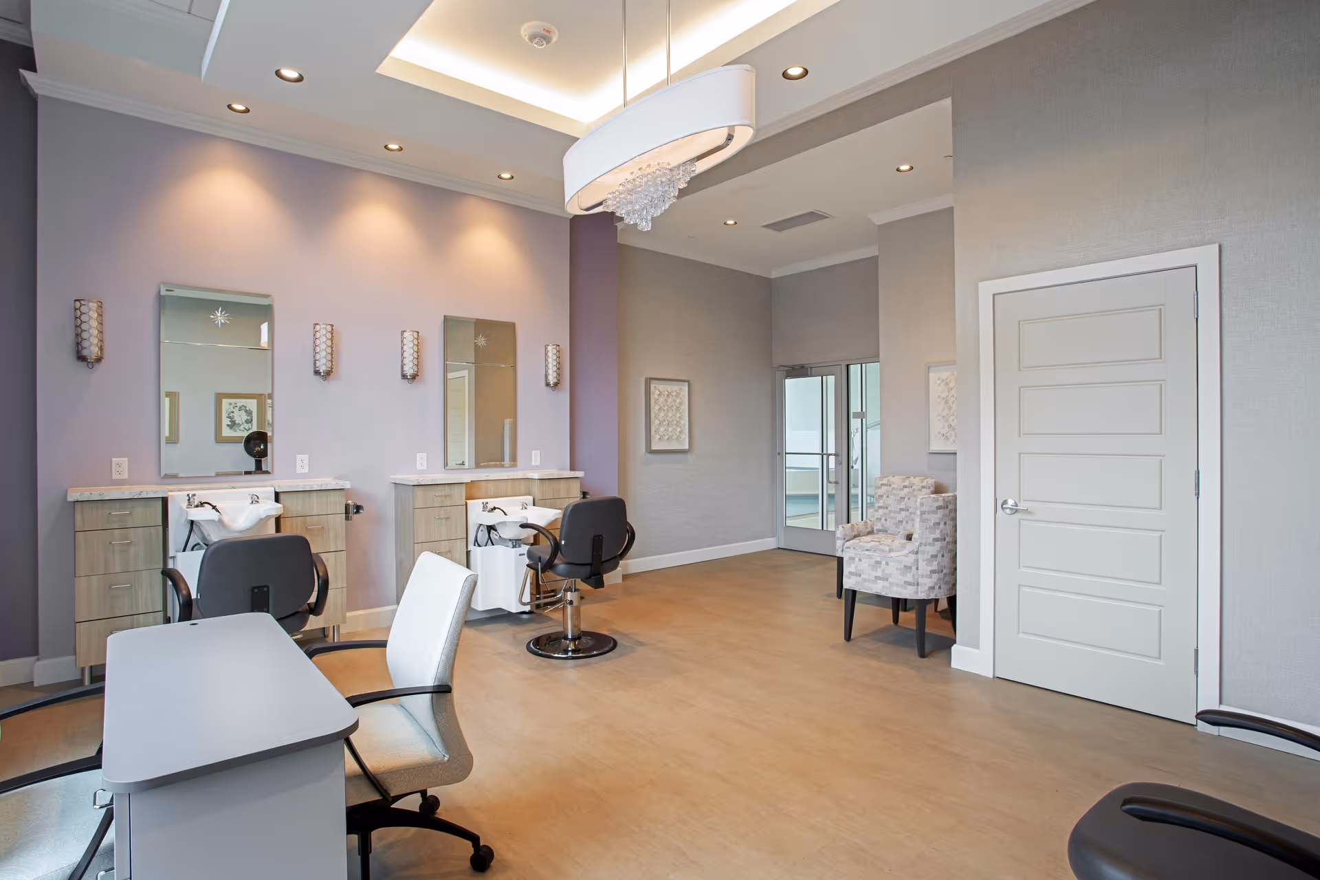 A modern salon area inside a senior living facility with two styling stations featuring mirrors, chairs, and sinks for hair washing. The room has light purple and gray walls, recessed lighting, a decorative ceiling light fixture, and a small seating area with two patterned armchairs near a door.