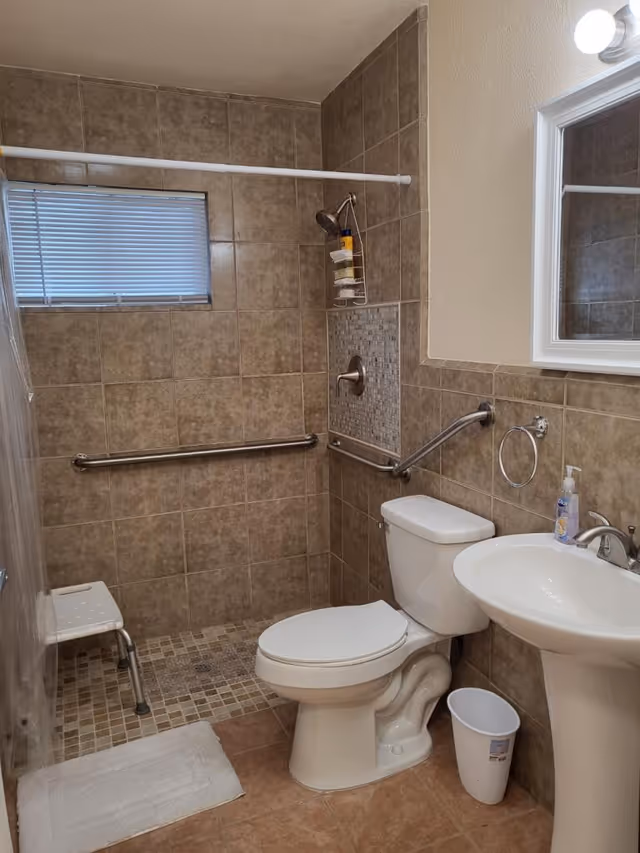 A bathroom with beige tiled walls and floor. It features a white toilet, a white pedestal sink with a soap dispenser, a small white trash bin, and a wall-mounted mirror with a light above it. There is a walk-in shower area with a handheld showerhead, grab bars, a small shower stool, and a window with closed blinds.