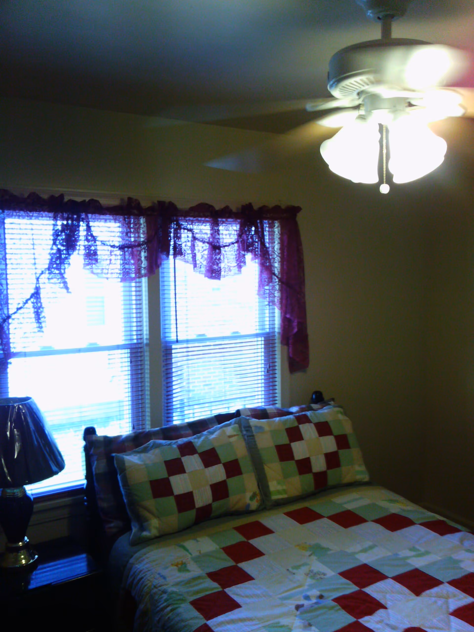 A cozy bedroom featuring a quilted bedspread with red and green patterns, a bedside lamp, and two windows with lace curtains.