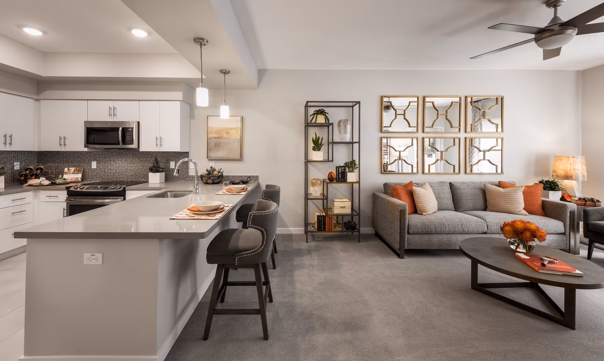 Modern open-concept living area featuring a kitchen with white cabinets, a gray countertop island with three bar stools, and a living room with a gray sofa, decorative pillows, a coffee table with a flower arrangement, a standing lamp, and a wall-mounted set of six decorative mirrors.