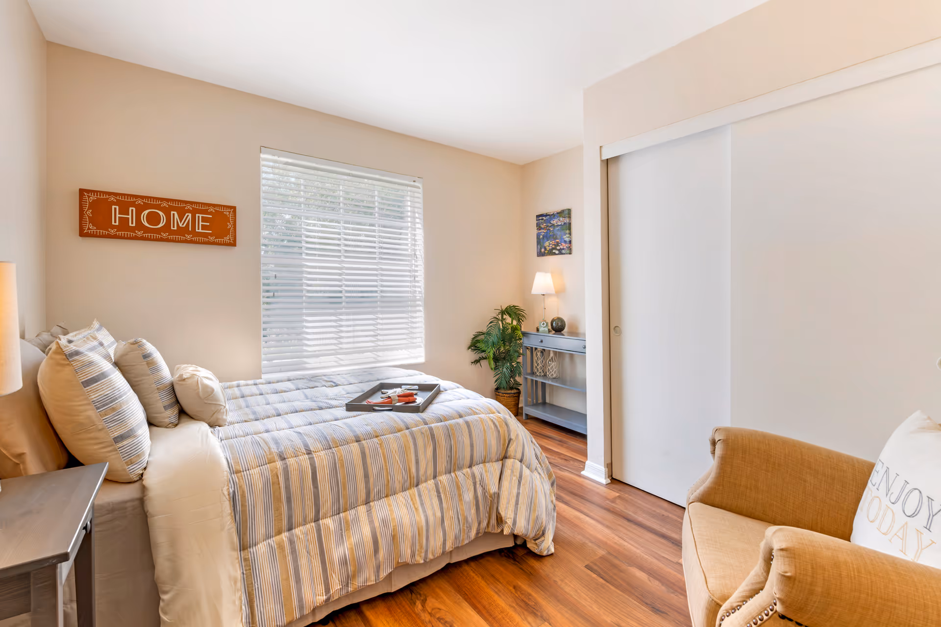 Sunlit cozy bedroom with a bed dressed in striped bedding, an armchair, window with blinds, and a 'HOME' wall sign.