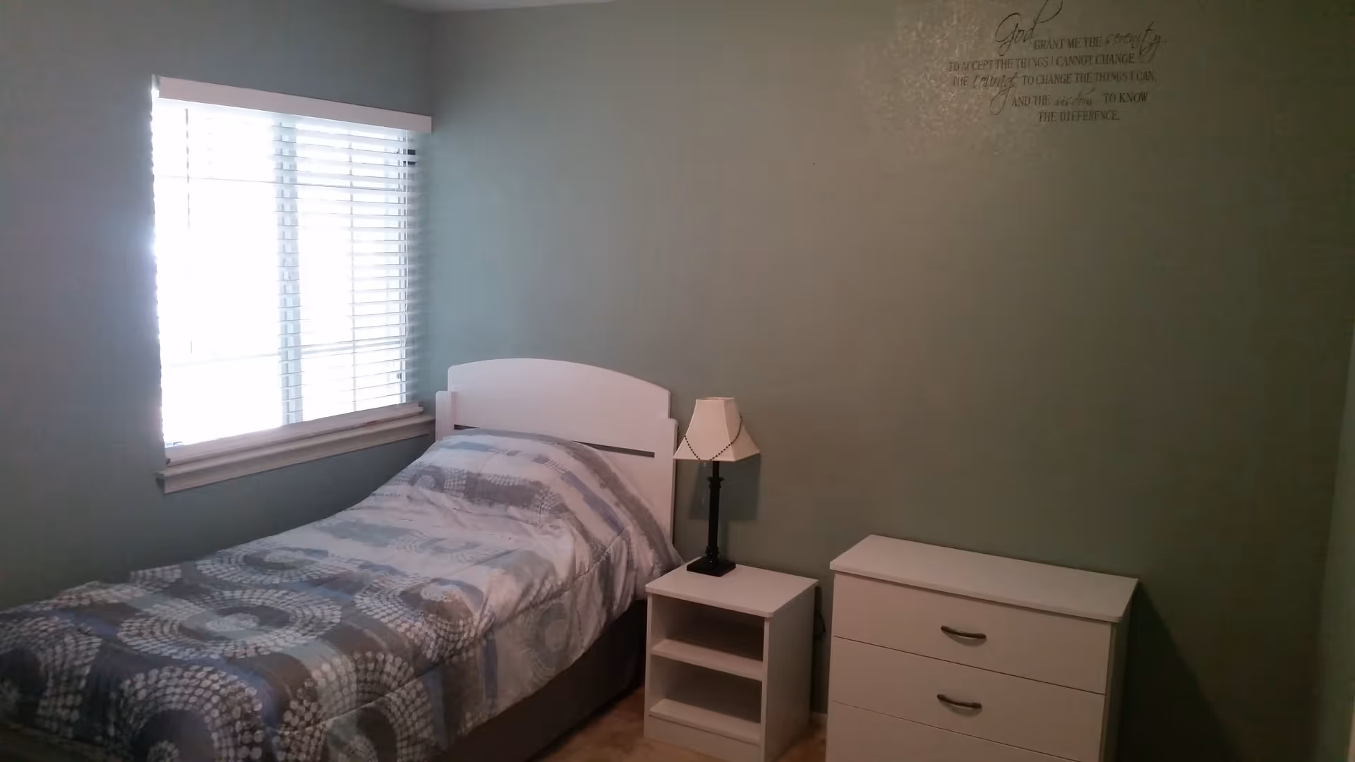 A small bedroom with a single bed covered with a patterned comforter, a white headboard, a white nightstand with a lamp, and a white dresser. There is a window with blinds on the left wall and a wall decal with a prayer quote on the right wall.