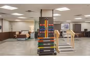 A spacious rehabilitation room with exercise equipment including colorful resistance bands on a rack and a set of stairs for physical therapy. The room has neutral-colored walls, ceiling lights, and some seating areas with chairs and a couch. There is a framed picture on the wall and a coat rack in the corner.