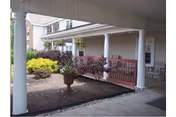 Covered outdoor porch area with white columns and a garden bed containing shrubs and a large planter. The porch has a railing and chairs visible near the entrance of a building.