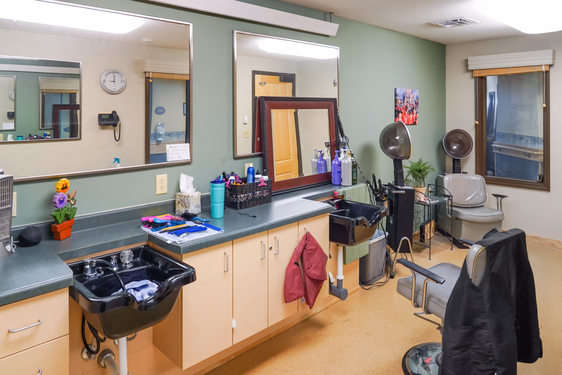 Interior view of a hair salon area in a senior living facility with two black hair washing sinks, salon chairs, hair dryers, large mirrors, and various hair care products on the counter. The room has light green walls and a window with a white blind.