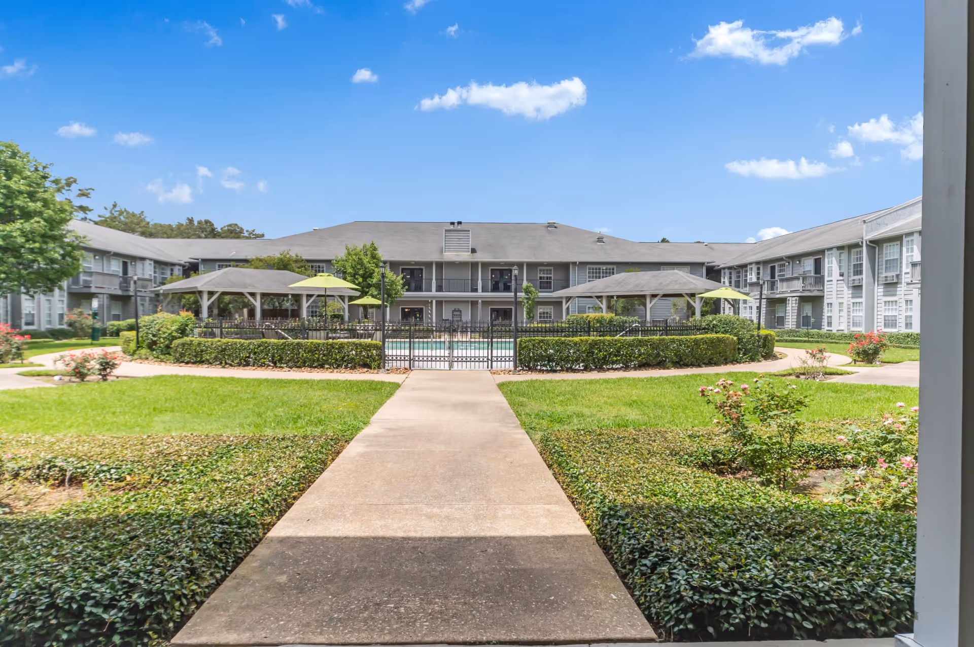 Central walkway leading to a gated pool and shaded pavilions in a landscaped courtyard surrounded by a two-story residential building under a blue sky.