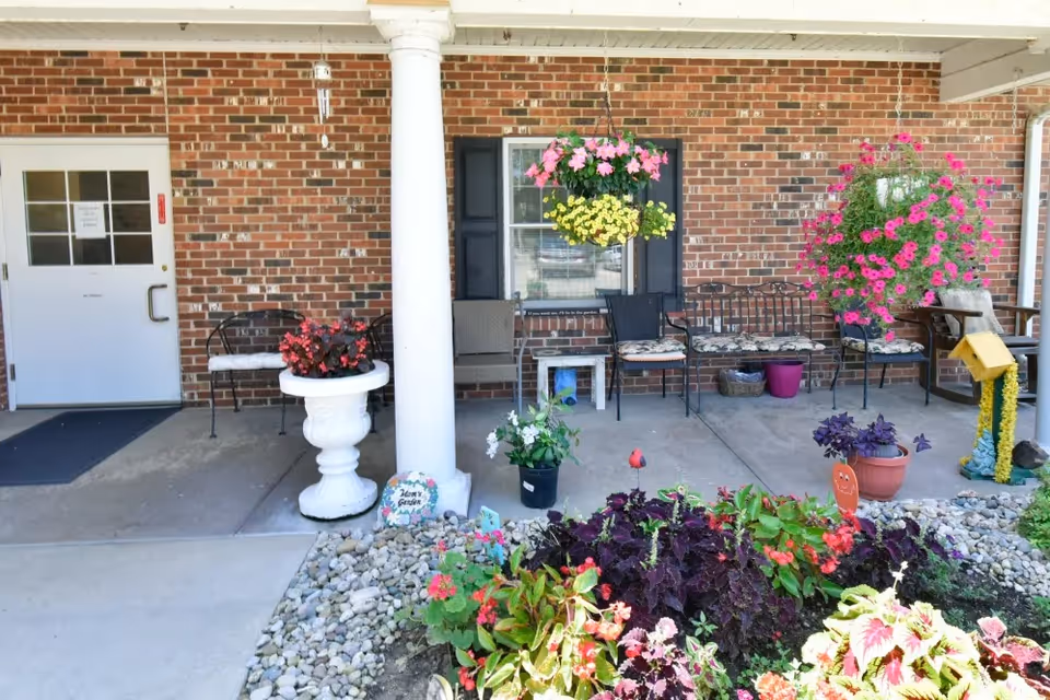 Outdoor patio area with a brick wall background, white column, hanging flower baskets, potted plants, benches with cushions, and a garden bed with various colorful flowers and rocks in front.