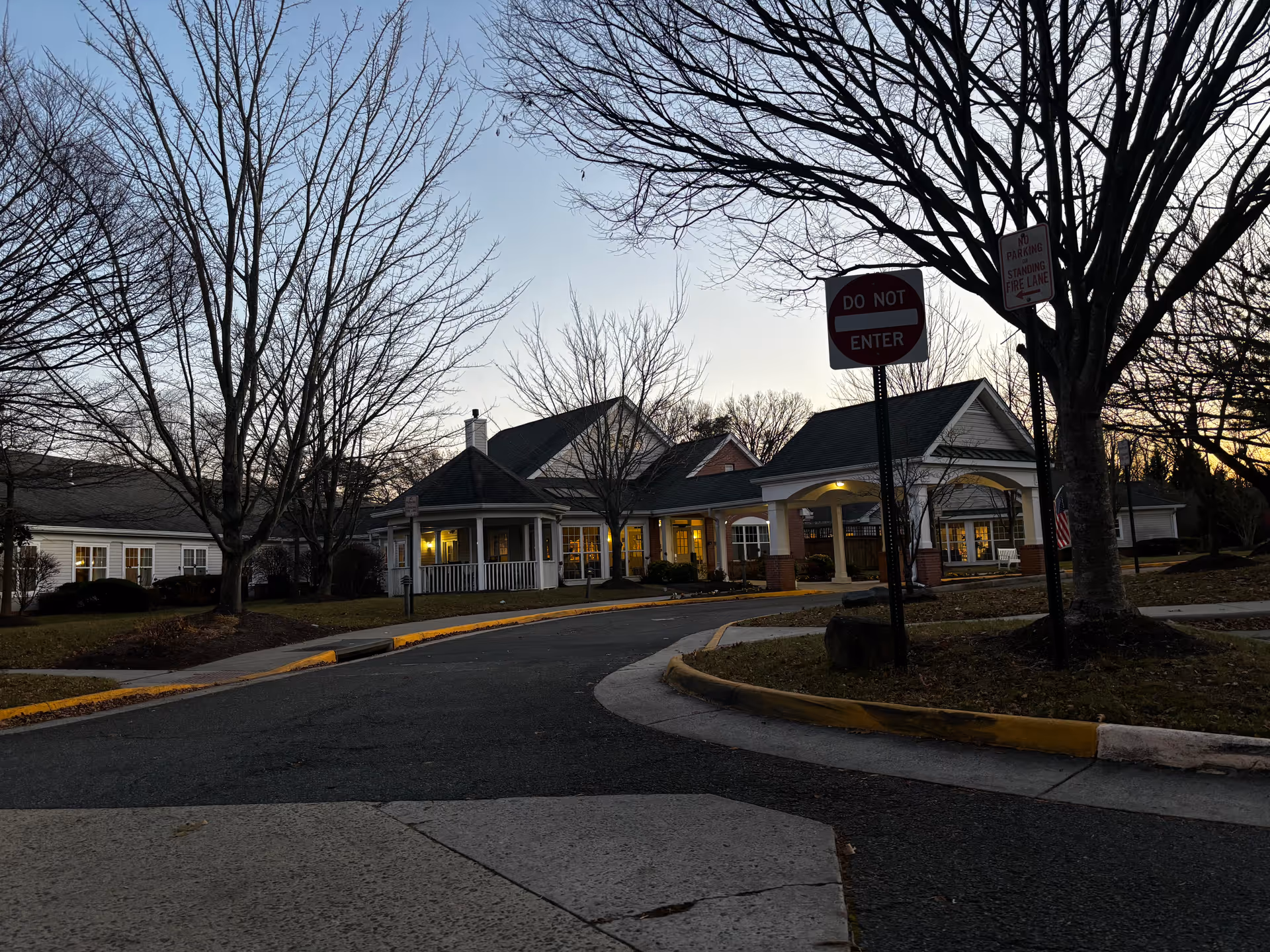 Front entrance of a senior living building at dusk with a covered porte-cochère, circular driveway, leafless trees, and a 'Do Not Enter' sign.