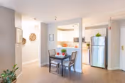 A bright and clean senior living facility interior featuring a small dining area with a table and two chairs. The dining area is adjacent to a kitchen with a stainless steel refrigerator, microwave, and light-colored cabinetry. The space has light wood flooring, neutral-colored walls, and decorative elements including plants and wall art.