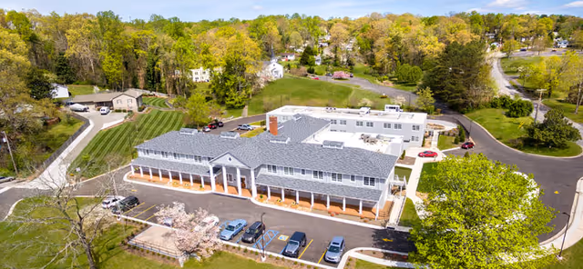 Aerial view of Alexander Guest House, a large two-story building with a gray roof and white columns along the front porch. The building is surrounded by green trees and lawns, with a parking lot in front containing several cars. A winding road leads to the facility, and additional smaller buildings and wooded areas are visible in the background under a clear blue sky.