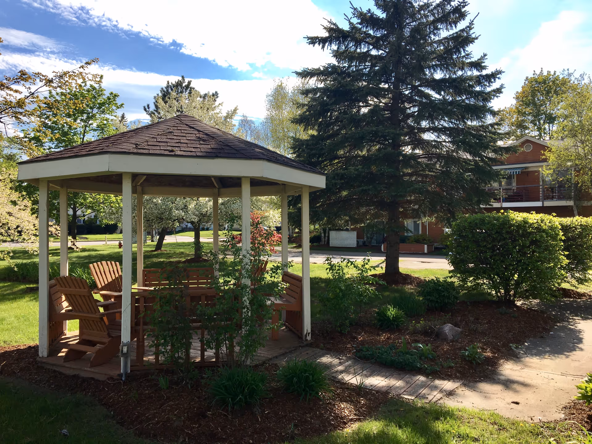 A wooden gazebo with Adirondack-style chairs sits on landscaped grounds with trees and an apartment building in the background.