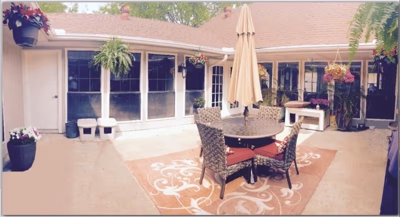 Outdoor patio area with a round table and four wicker chairs with red cushions, centered on a decorative outdoor rug. A large beige umbrella is positioned in the middle of the table. The patio is surrounded by a building with multiple windows and a door. Various hanging and potted plants decorate the space, creating a cozy and inviting atmosphere.