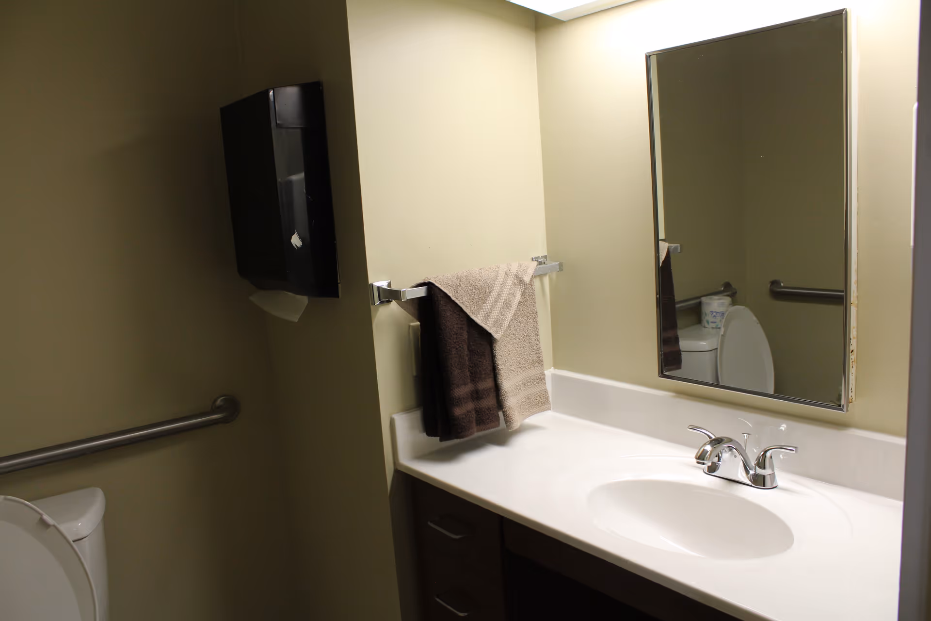 A bathroom with a white sink and countertop, a silver faucet, a rectangular mirror above the sink, a towel rack with two towels (one beige and one brown), a toilet with the lid up, a grab bar on the wall, and a black paper towel dispenser mounted on the wall.