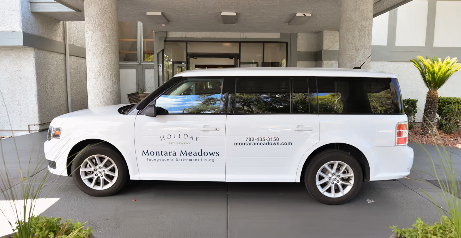 A white shuttle vehicle parked under a covered entrance of a building. The vehicle has signage on the side that reads 'Holiday Retirement Montara Meadows Independent Retirement Living' along with a phone number and website. There are plants and a palm tree near the entrance.